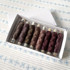 May include: A box containing eight spools of brown embroidery thread. The spools are labelled with numbers and the word "farbo".