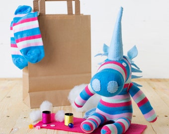 May include: A blue and pink striped unicorn stuffed animal with a brown paper bag and blue and white striped socks. The unicorn is made from fabric scraps and has a long, pointed horn. There are also two spools of thread and some cotton balls on a pink surface.