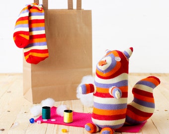 May include: A striped sock puppet in the shape of a squirrel with a brown paper bag and a sock in the background.