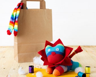 May include: A colorful sock lion with a red body, blue eyes, and a rainbow striped mane. The lion is sitting on a yellow felt surface with white stuffing and spools of thread nearby.