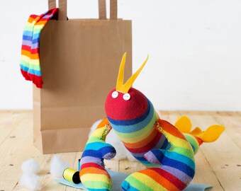 May include: A rainbow striped sock lobster toy with yellow antennae and button eyes. The toy is sitting on a light blue surface with white cotton balls and a brown paper bag in the background.