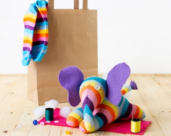 May include: A colorful striped sock elephant toy with purple ears and googly eyes. The elephant is lying on a red fabric with white stuffing and spools of thread nearby.