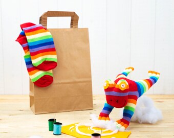 May include: A rainbow striped sock frog toy with a brown paper bag and spools of thread on a wooden surface.