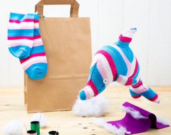 May include: A pink and blue striped dolphin plush toy with a brown paper bag and a pair of pink and blue striped socks. The dolphin is flying in the air and the bag is on a wooden surface.