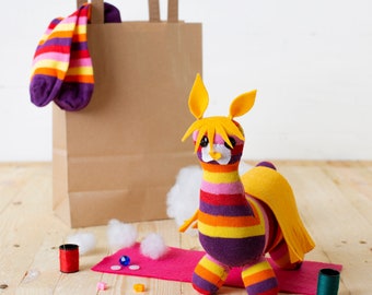 May include: A colorful striped llama toy made from recycled socks. The llama has a yellow head and a purple body with orange, yellow, and pink stripes. The llama is sitting on a pink felt mat with white stuffing and a brown paper bag in the background.