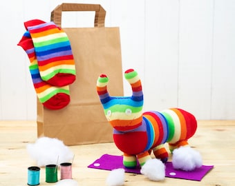 May include: A handmade sock creature with rainbow stripes, red accents, and button eyes, sits on a purple mat. A pair of rainbow striped socks and a brown paper bag are in the background. Sewing supplies and cotton balls are also present.