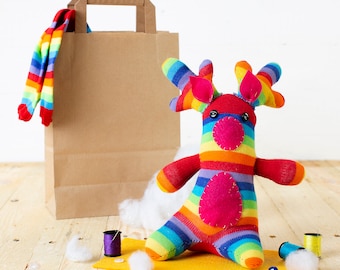 May include: A colorful rainbow striped reindeer toy made from recycled socks. The reindeer has a pink nose and is sitting on a yellow surface with white stuffing and spools of thread.