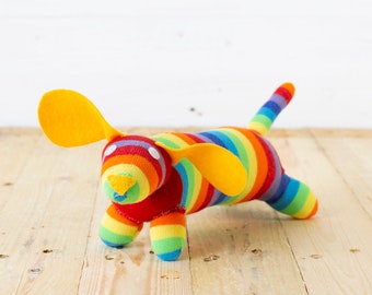 May include: A plush dog toy crafted from colorful striped fabric featuring a rainbow pattern. The toy has two large, solid yellow felt ears, a small yellow felt nose, and two white circular eyes. Its body, legs, and tail are covered in horizontal stripes of red, orange, yellow, green, blue, and purple.