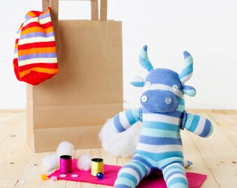 May include: A blue and white striped sock monkey with a brown paper bag and a red and white striped sock in the background. The monkey is sitting on a pink felt mat with two spools of thread and cotton balls.