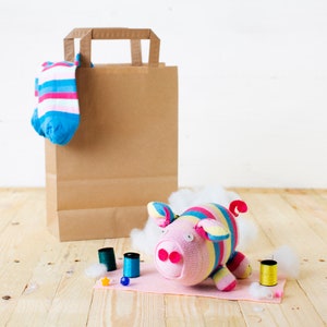 May include: A pink and rainbow striped sock monkey with a brown paper bag and a sock in the background. The monkey is sitting on a pink surface with white cotton balls and spools of thread.