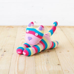 May include: A stuffed cat toy crafted from multi-coloured striped fabric in shades of pink, yellow, and blue. The toy features two white button eyes, a stitched triangular nose, and small purple fabric ears, with a long, upright tail.