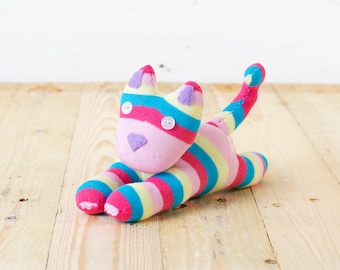 May include: A stuffed cat toy crafted from multi-colored striped fabric in shades of pink, yellow, and blue. The toy features two white button eyes, a stitched triangular nose, and small purple fabric ears, with a long, upright tail.