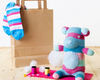 May include: A striped sock teddy bear with pink and blue stripes, white stuffing, and black eyes. The bear is sitting on a pink fabric with white cotton balls and spools of thread nearby. A brown paper bag with a striped sock hanging out of the top is in the background.
