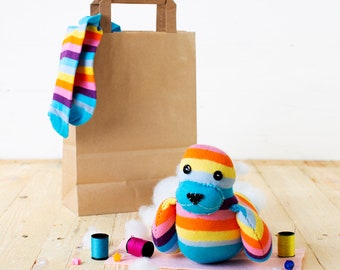 May include: A colorful striped sock monkey toy with blue eyes and a brown nose. The monkey is sitting on a white fluffy surface next to a brown paper bag with a sock sticking out of the top. There are also two spools of thread and a few small beads on the surface.