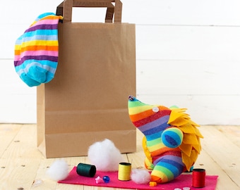 May include: A handmade sock hedgehog made from a colorful striped sock, with yellow felt spines and button details. A brown paper bag and a striped sock hang in the background. Sewing supplies, including thread and stuffing, are arranged on a pink felt square.