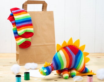 May include: A rainbow striped sock dinosaur toy with yellow spikes, a brown paper bag, and two rainbow striped socks. There are also white cotton balls, spools of thread, and a piece of paper on a wooden surface.