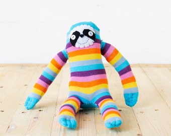 May include: A stuffed sloth toy crafted from multi-colored striped fabric featuring horizontal bands of blue, orange, yellow, purple, and pink. The face has a white circular patch with black eye markings, small white button eyes, and a stitched mouth.