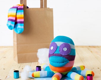 May include: A colorful octopus made from fabric scraps with purple and white buttons for eyes. The octopus is sitting on a wooden surface with a brown paper bag and spools of thread nearby.