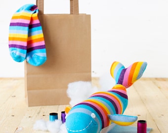 May include: A colorful whale stuffed animal made from striped socks. The whale is blue with a white belly and has a rainbow striped tail and fins. The whale is sitting on a white fluffy surface with a brown paper bag and a pair of striped socks in the background.