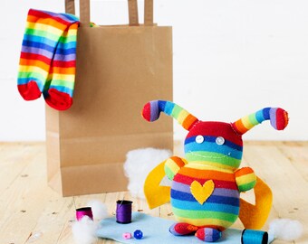 May include: A rainbow striped stuffed toy with a heart on its chest. The toy has wings and antennae. The toy is sitting on a blue surface with spools of thread and beads.