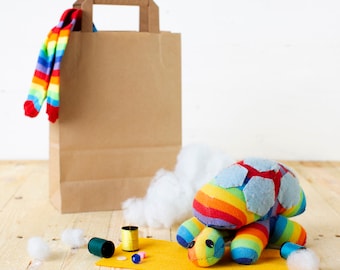 May include: A rainbow-colored sock turtle toy with blue felt accents, a brown paper bag, and white stuffing. The toy is on a yellow surface with spools of thread and buttons.