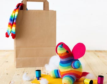 May include: A handmade sock animal in rainbow colors with pink ears and a red tail sits on a yellow felt square. A brown paper bag and a pair of rainbow socks are in the background. Sewing supplies, including thread and cotton, are scattered around.