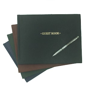 Könnte beinhalten: Ein Stapel aus vier dunkelgrünen, braunen und blauen Gästebüchern mit einem silbernen Stift darauf. Das oberste Buch trägt die Aufschrift "GUEST BOOK" in goldenen Buchstaben.