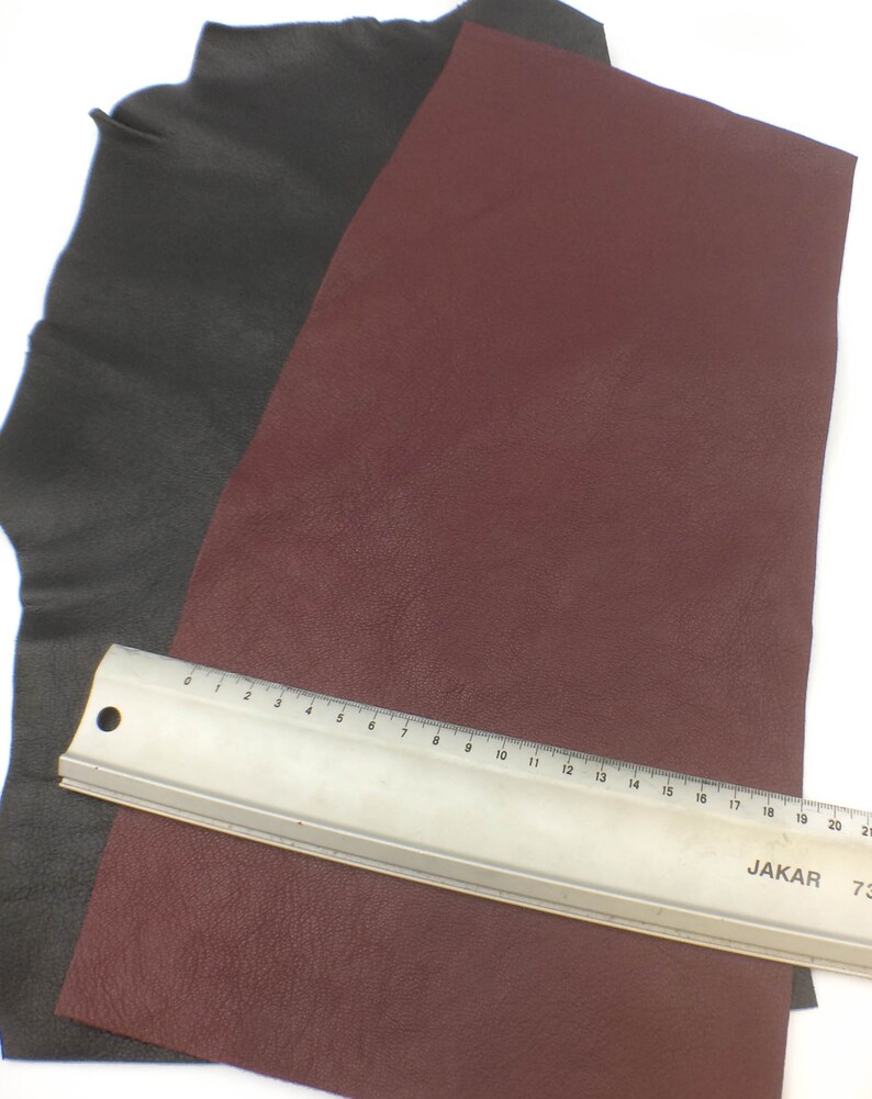 May include: Two pieces of leather, one black and one burgundy, are laid out on a white surface. A metal ruler with markings in inches is placed on top of the burgundy leather.