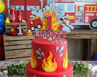 May include: A two-tiered red birthday cake decorated with flames, water splashes, and a topper that reads "ELLIOTT'S 1ST BIRTHDAY! FIRE PATROL IN SERVICE!". The cake is on a white plate, with Dalmatian figurines and fire truck cutouts nearby.