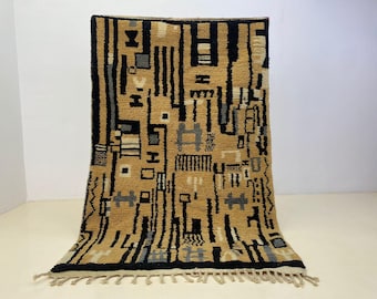 May include: A rectangular area rug featuring a tan background with a complex geometric pattern in black, grey, and cream. The design consists of vertical lines, squares, and abstract shapes. The rug has a thick, textured pile and a fringed edge.