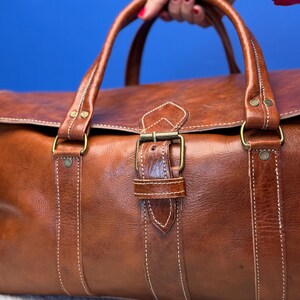 以下が含まれることがあります： バックル留めと白いステッチが付いた茶色の革製のダッフルバッグ。バッグには2つのトップハンドルが付いています。