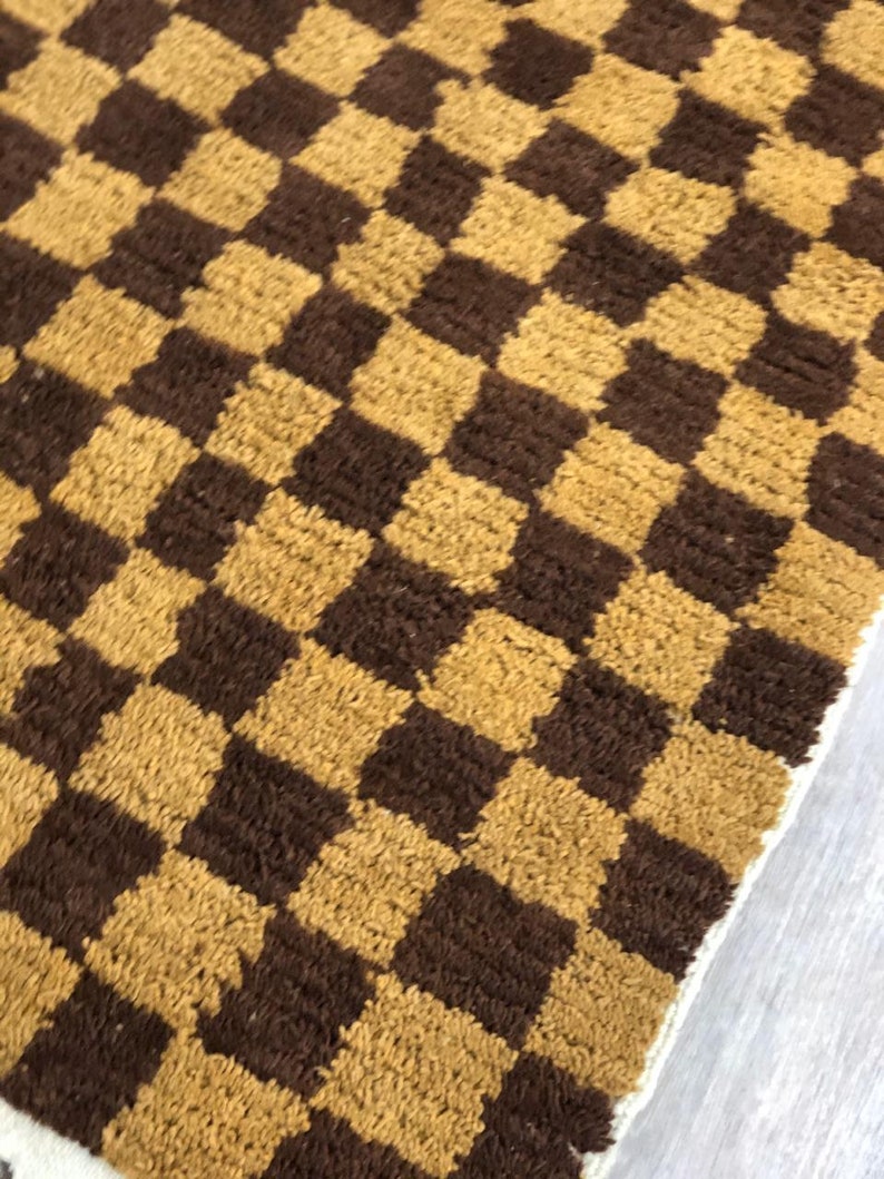 K&ouml;nnte beinhalten: Ein brauner und goldener karierter Teppich mit einem wei&szlig;en Fransenrand.