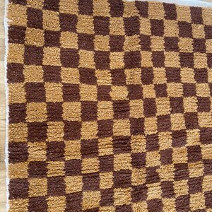 K&ouml;nnte beinhalten: Ein brauner und goldener karierter Teppich mit wei&szlig;em Rand. Der Teppich ist aus einem weichen, flauschigen Material.