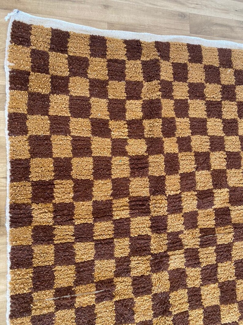 K&ouml;nnte beinhalten: Ein brauner und goldener karierter Teppich mit wei&szlig;em Rand. Der Teppich ist aus einem weichen, flauschigen Material.