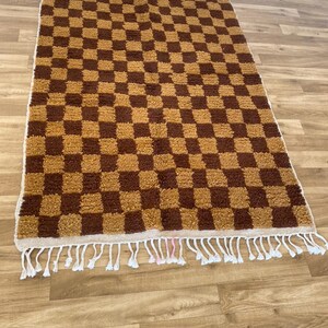K&ouml;nnte beinhalten: Ein brauner und beigefarbener Schachbrettteppich mit einem wei&szlig;en Fransenrand. Der Teppich hat eine weiche, flauschige Textur.