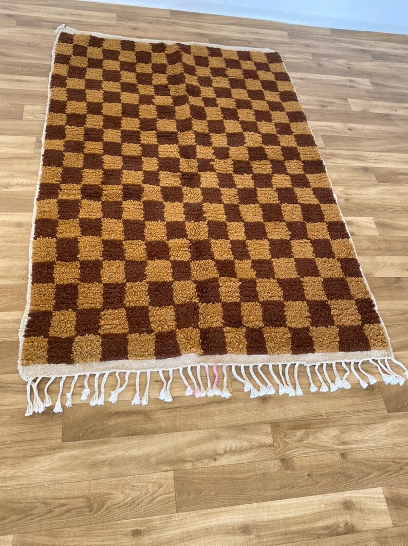 K&ouml;nnte beinhalten: Ein brauner und beigefarbener Schachbrettteppich mit einem wei&szlig;en Fransenrand. Der Teppich hat eine weiche, flauschige Textur.