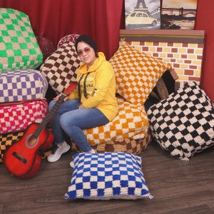 Puede incluir: Una mujer se sienta en una pila de cojines de suelo a cuadros de colores, sosteniendo una guitarra acústica roja. Los cojines están hechos de un material suave y esponjoso y están dispuestos en forma de pirámide. La mujer lleva una sudadera con capucha amarilla y pantalones vaqueros azules.