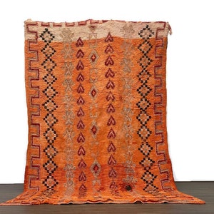 May include: A vintage Moroccan rug with a geometric pattern in shades of orange, red, and black. The rug has a fringed edge and is made of wool.