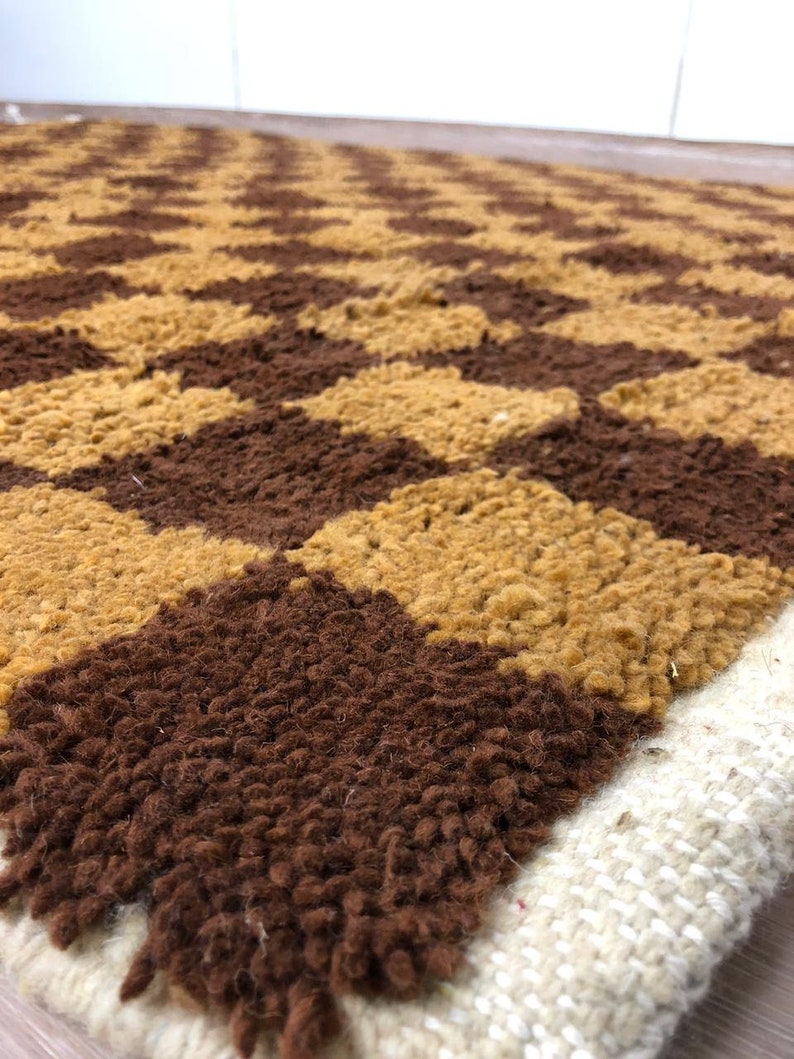 K&ouml;nnte beinhalten: Ein brauner und goldener geometrisch gemusterter Teppich mit einem wei&szlig;en Fransenrand. Der Teppich ist aus einem weichen, flauschigen Material.