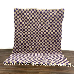 May include: A checkered rug with a beige and purple pattern. The rug has a fringe border.