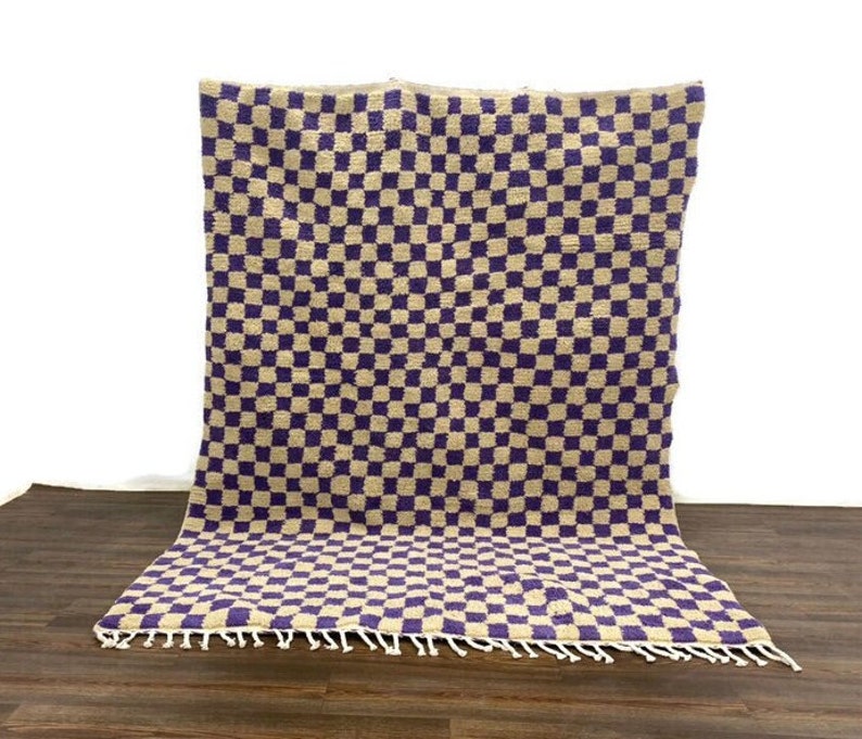 May include: A checkered rug with a beige and purple pattern. The rug has a fringe border.
