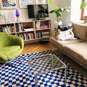 May include: A blue and white checkered rug with a glass coffee table in the center. A green armchair is in the foreground, and a couch with a throw blanket is in the background.