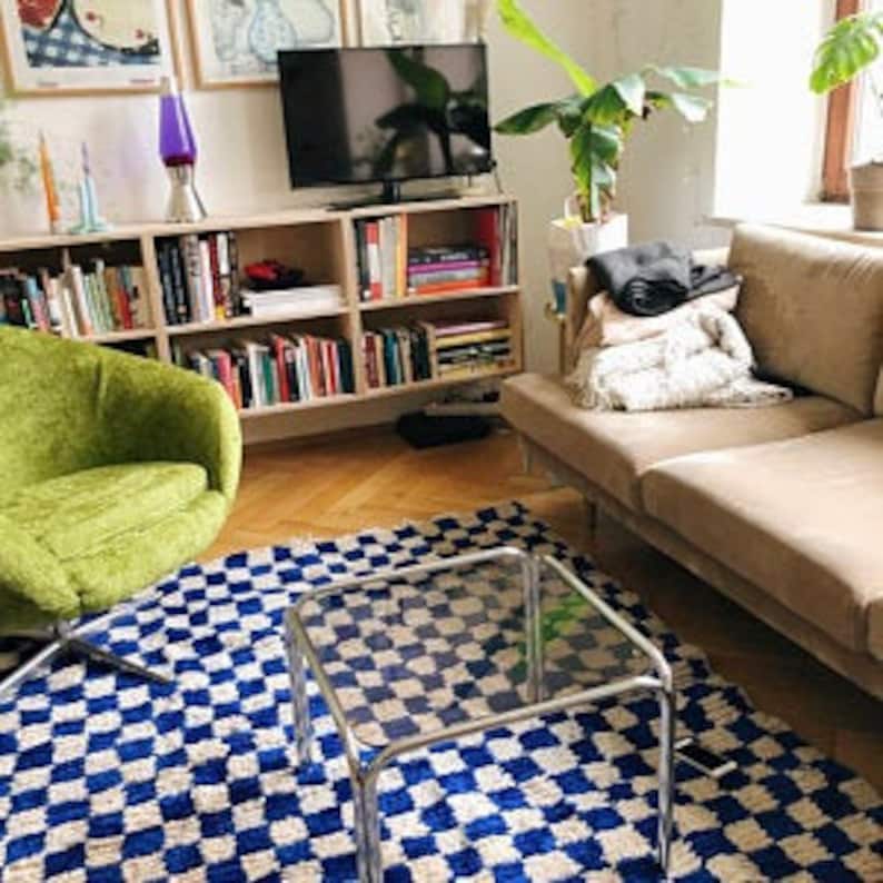 May include: A blue and white checkered rug with a glass coffee table in the center. A green armchair is in the foreground, and a couch with a throw blanket is in the background.