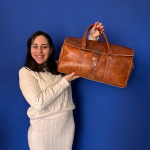 以下が含まれることがあります： 女性が真鍮のバックルが付いた茶色の革製のダッフルバッグを持っています。バッグはトップハンドルとショルダーストラップが付いています。女性は白いケーブルニットのセーターとスカートを着用しています。