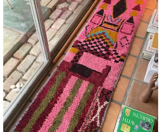 May include: A long, pink and burgundy runner rug with a geometric pattern in shades of brown, yellow, and blue. The rug has a fringe on both ends.