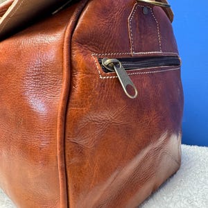 以下が含まれることがあります： 真鍮製のハンドルとジッパー付きポケットが付いた、茶色の革製のダッフルバッグ。バッグの上部に折り畳んでバックルで固定できるフラップが付いています。