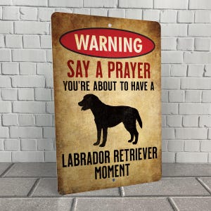 May include: A rectangular metal sign with a distressed, vintage look. The sign features the text "WARNING SAY A PRAYER YOU'RE ABOUT TO HAVE A LABRADOR RETRIEVER MOMENT" in red and brown lettering, with a black silhouette of a Labrador Retriever.