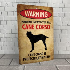 May include: A warning sign with a black silhouette of a Cane Corso dog. The sign reads "WARNING PROPERTY IS PROTECTED BY A CANE CORSO CANE CORSO IS PROTECTED BY MY GUN".