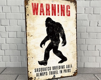 May include: A vintage-style warning sign with a distressed background and rusty edges. The sign features a black silhouette of a Sasquatch walking and the text "WARNING" in red letters. The text "Sasquatch Breeding Area Always Travel in Pairs" is printed in black at the bottom of the sign.