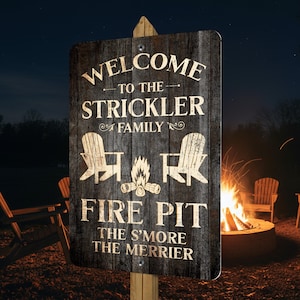 May include: A dark wooden sign with the text "WELCOME TO THE STRICKLER FAMILY FIRE PIT THE S'MORE THE MERRIER." The sign features illustrations of Adirondack chairs and a campfire. The background shows a fire pit with a lit fire and Adirondack chairs.